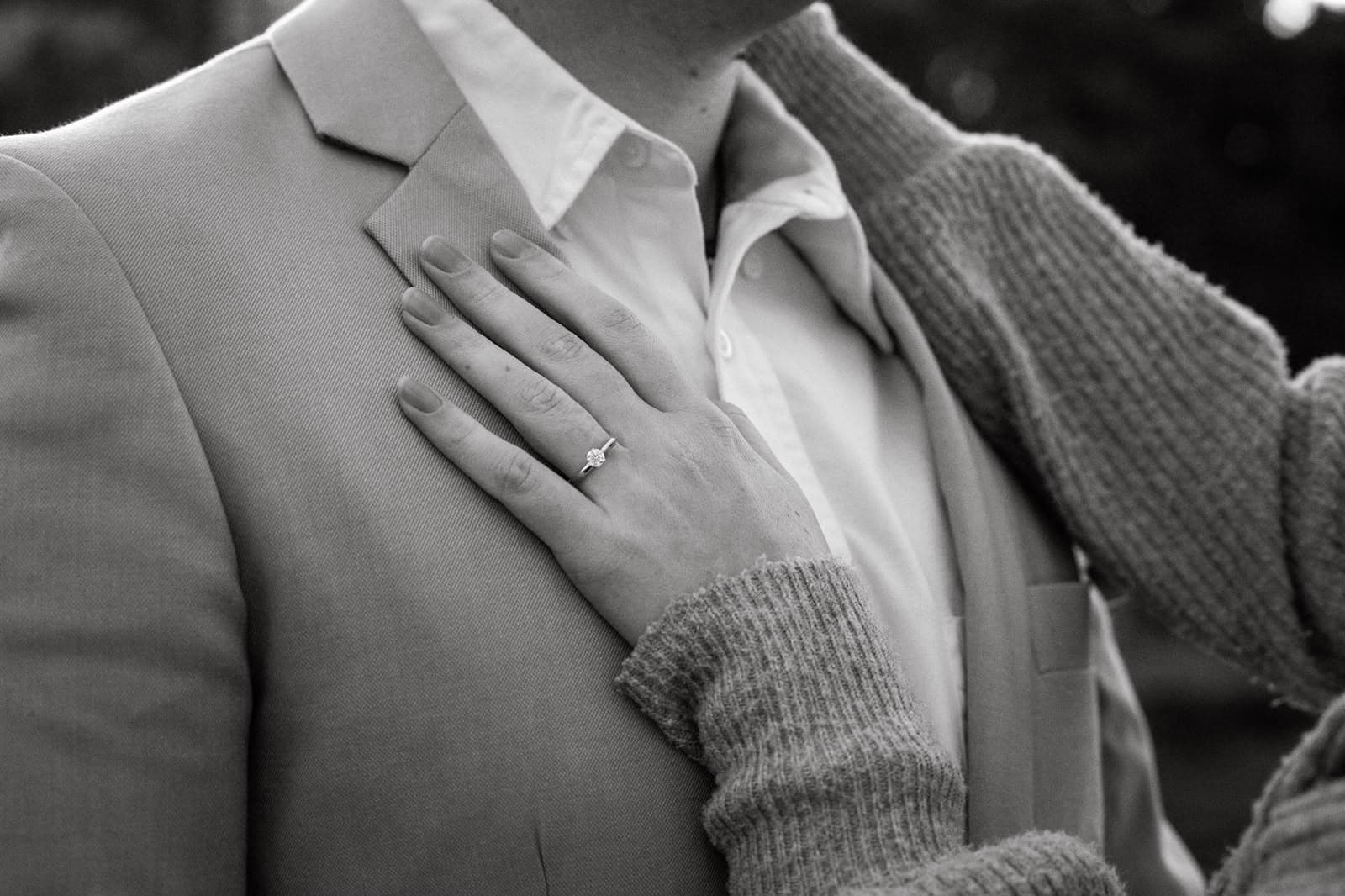 Hand with a diamond engagement ring resting on a suit jacket in black and white.